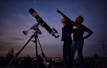 Silhouettes of father, daughter and astronomical telescope under starry skies.