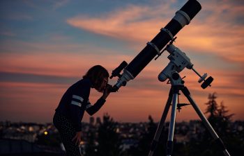 Girl with astronomical telescope stargazing under twilight sky.